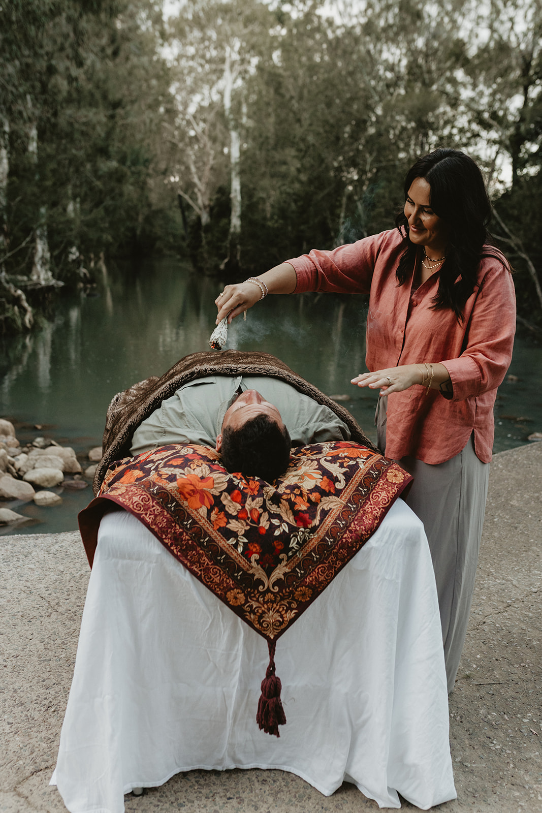 Charlene using a sage bundle for energy clearing during an outdoor healing session beside the water in Sarina QLD, creating a calm and supportive space for release.