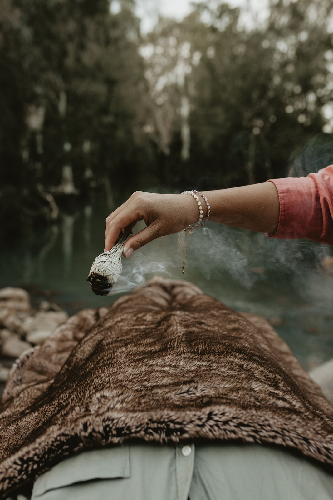 Close-up of Charlene holding burning sage over a client wrapped in a blanket during an energy healing session by a tranquil creek in Sarina QLD.