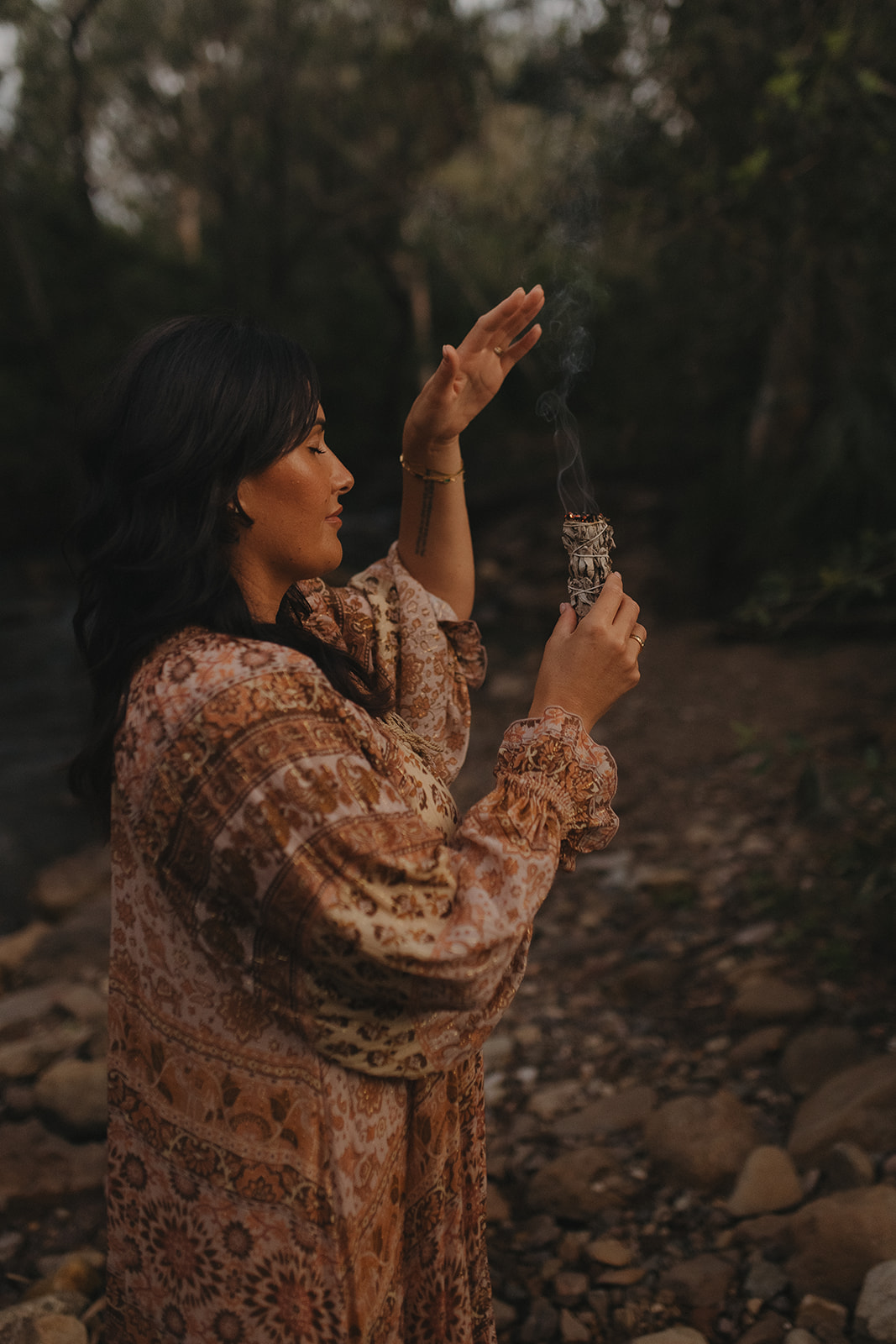 Charlene seated on river stones in a white flowing dress, gently playing a frame drum during a sacred sound healing session on her rural property in Sarina QLD.